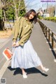 A woman in a green jacket and blue pleated skirt is walking down the street.